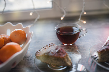 transparent glass tea mug stands on a wooden table with cake and tangerines in the Christmas lights