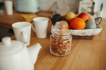 white mugs of tea and coffee with a transparent glass jar of brown cane sugar stand on a wooden table on a background of fruits