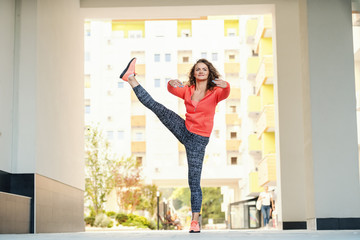Good-looking smiling caucasian woman in sportswear and with curly hair doing side high kicks while standing in passage on sunny day.