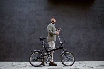 Fototapeta na wymiar Full length of smiling caucasian fashionable man pushing bicycle and holding smart phone while passing by gray wall.
