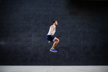 Side view of handsome muscular caucasian man in shorts and t-shirt skipping rope in front of gray wall outdoors.