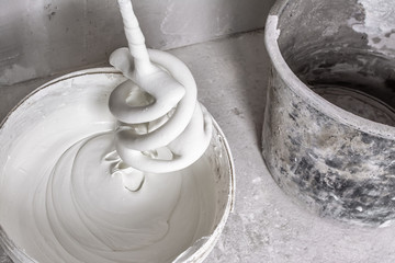 Plastic bucket with fresh putty, spiral whisk for electric building mixer soiled with putty and empty dirty bucket on raw concrete floor. Facing work and construction concept. Close-up