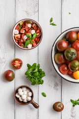 Ceramic bowl with caprese salad, fresh black prince tomatoes and yellow tomatoes in enamel pan, mozzarella cheese and basil leaves on white wooden table. Organic and vegetarian food concept. Vertical