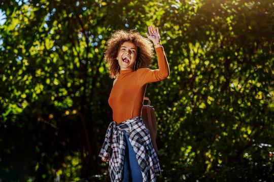 Attractive young mixed race woman dressed casual and with curly hair standing in park and waving to a friend.