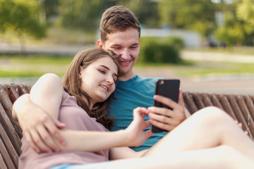 Young couple  look at the smartphone screen during a summer walk sitting on a bench