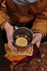  metal cup of tea with lemon and ginger in the hands of a man in a yellow jacket, on a wooden plate and with autumn leaves