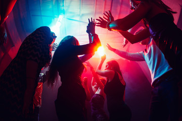 Until sunrise. A crowd of people in silhouette raises their hands on dancefloor on neon light...