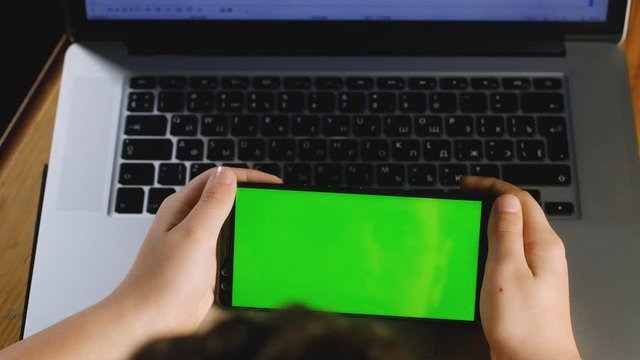 child is sitting at table with laptop looking at smartphone with green screen. concept of viewing photos and videos on a smartphone. Suitable for simulation of watching the news on phone. chroma key.