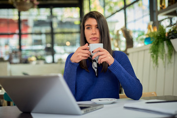 Attractive caucasian in sweater sitting in cafe, holding coffee. On table is laptop.