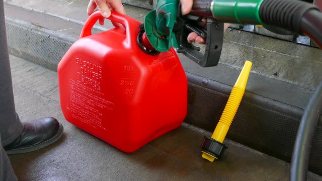 Close POV shot of a woman&rsquo;s hands using a fuel pump nozzle to fill a red plastic fuel container with unleaded petrol / gas, at a gas / petrol station.