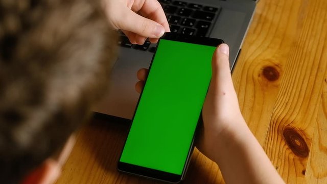 child is sitting at table with laptop looking at smartphone with green screen. Your finger makes scrolling motion. Suitable for simulating news viewing apps photos and videos on your phone. chroma key