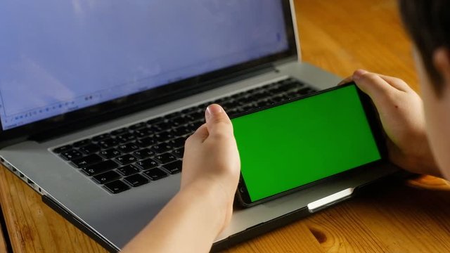 child sits at a table with a laptop holding a smartphone with a green screen with two hands. Suitable for simulating news viewing apps photos and videos on your phone. the chroma key.