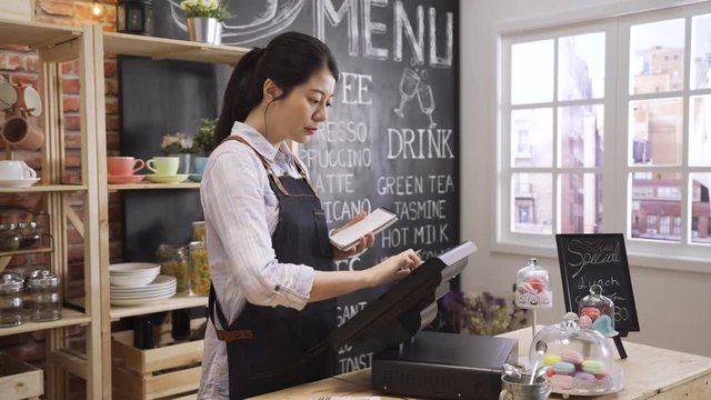 Young Asian korean girl waitress uniform making notes in notebook and typing on digital tablet on counter table. cafe female worker in apron enter customer order in point of sale terminal machine