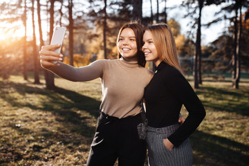 Happy two young twin sisters in casual outfit with bright smile take selfie with smartphone at the park