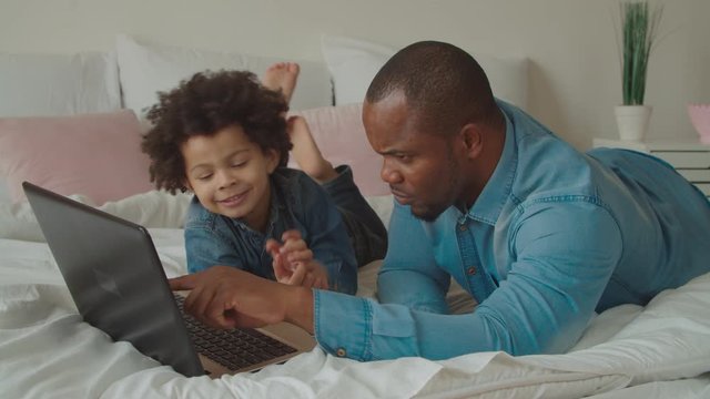 Positive caring handsome african father and joyful adorable preschooler mixed race son networking with laptop pc on bed, communicating ,watching online educational and developing programs for kids.