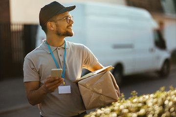 Happy delivery man text messaging on mobile phone while carrying packages. - obrazy, fototapety, plakaty