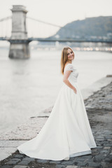 Fototapeta na wymiar Beautiful brunette bride in elegant white dress holding bouquet posing near river
