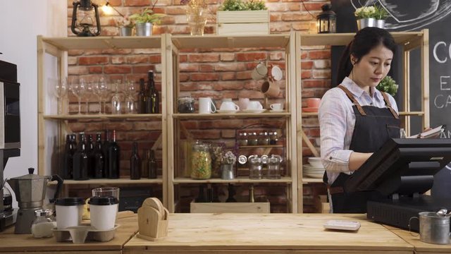 charming asian korean woman waitress in apron standing in counter bar working preparing customer order. attractive lady barista holding bread tong putting crispy croissant on plate in coffee shop.