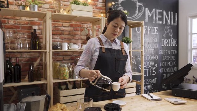 attractive asian female barista pouring black coffee in cup in counter bar. elegant chinese girl waitress making tasty drink for customer in cafe store. coffeehouse worker holding white cup.