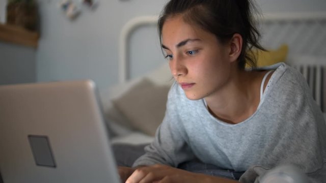 Close up of mixed-race teenage girl lying in bed in her bedroom, looking at laptop screen and typing messages to her friends