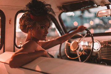 Portrait of beautiful sexy fashion blond girl. Model with bright makeup and curly hairstyle in retro style hat sitting in old car. Attractive woman  rides around the night city
