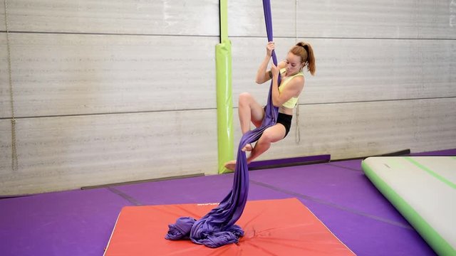 Young redheaded woman doing aerial gymnastic acrobatics with acrobatic fabrics inside a gym.