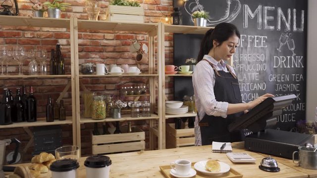 young female bartender prepared customer order and ringing bell calling staff waiter at coffee shop. asian chinese woman barista holding note writing and working on digital tablet in counter bar.