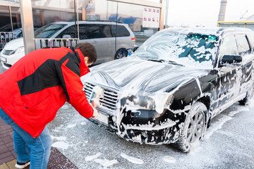A man in a red jacket wipes a foam-covered black car with a brush at a self-service car wash. Front view