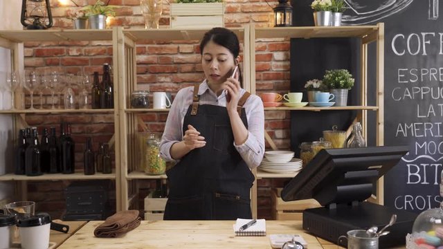beautiful smiling asian female barista taking order on cellphone call and writing note in cafeteria. young girl waitress listening to customer talking on mobile phone in counter bar of coffee shop.
