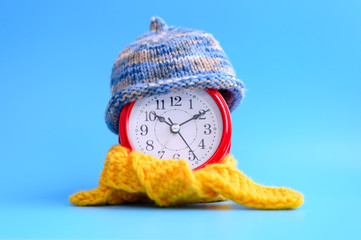 red round alarm clock in knitted wool blue hat and yellow scarf centered on a blue background....