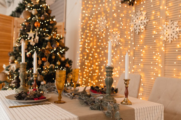 Served table with christmas decor and bokeh lights background, candles and christmas tree. New year celebration concept. 