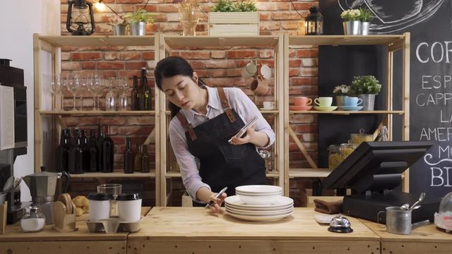 Pensive asian female waiter in apron counting cups and plates standing at bar counter during working process. woman employee of coffee shop making inventory to do report. lady barista making note.