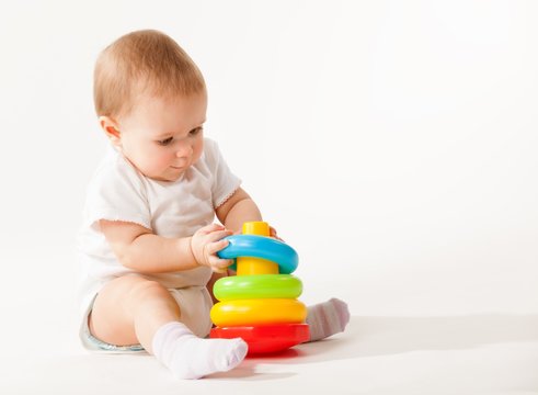 Charming cute baby in white overalls holds rings of a colored plastic pyramid sitting on a white background. Concept of smart and healthy little children concept of child care. Advertising space