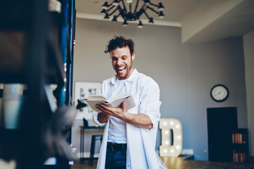 Happy excited hipster guy reading funny literature bestseller with funny jokes and laughing in home...