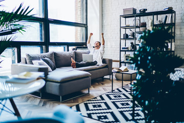 Overjoyed hipster guy feeling excited from win in online game on website spending weekend time in home interior with modern netbook, happy accomplished man resting on sofa and celebrating victory