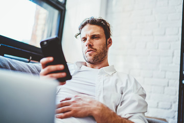 Fototapeta premium Pensive caucasian male millennial using mobile phone for read email notification while resting on sofa at home interior, serious hipster guy checking banking balance and payment via app on smartphone.