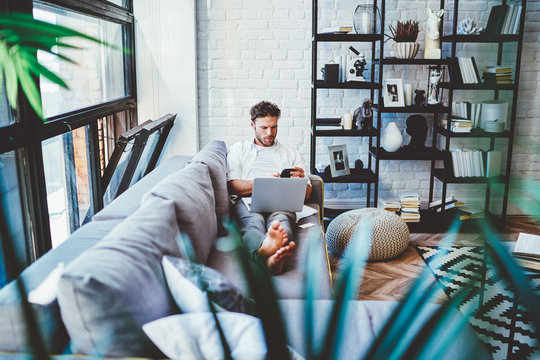Serious caucasian man using laptop computer for paying and shopping online via wifi, hipster guy lying on sofa in living room with netbook watching videos during free time at home interior.