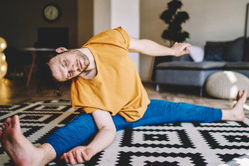 Young man with closed eyes sitting in twine on carpet and doing stretching exercises ro develop flexibility at home interior.Motivated male lover of yoga doing morning training in apartment