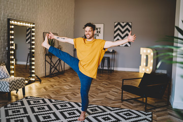 Happy young man holding leg in hand and standing on carpet doing stretching exercises during morning training in home apartment.Positive male lover of yoga practising meditation in different poses