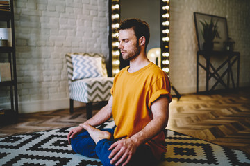Calm young man with closed eyes sitting in lotus pose on comfortable carpet meditating in morning...