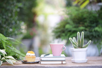 Pink coffee cup with cupcake  and small cactus plant and diary notebook on wooden table at outdoor