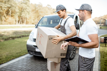 Two male couriers delivering goods by cargo van vehicle, unloading cardboard parcels from a car and checking list outdoors