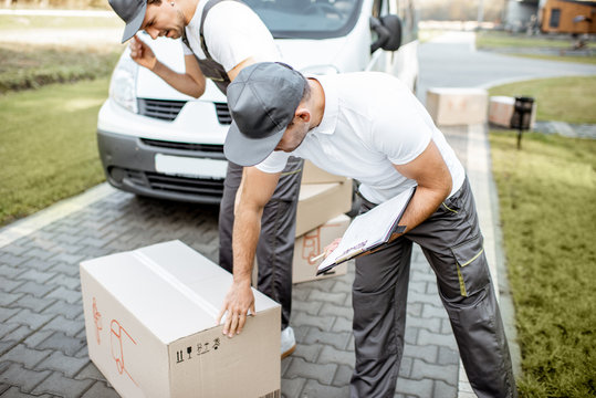 Two male couriers delivering goods by cargo van vehicle, unloading cardboard parcels from a car and checking list outdoors