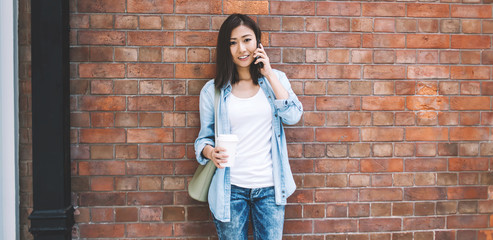 Asian hipster girl talking with friend via modern electronic gadget while standing near brick wall and looking at camera, young positive female teenager communicated in roaming using smartphone