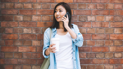 Young female person talking on mobile standing on urban brick wall background, attractive Chinese hipster girl having cellphone conversation in roaming near copy space for advertising text