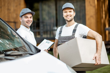 Portrait of a two cheerful delivery men in uniform standing together with check list and cardboard boxes near the cargo van vehicle outdoors