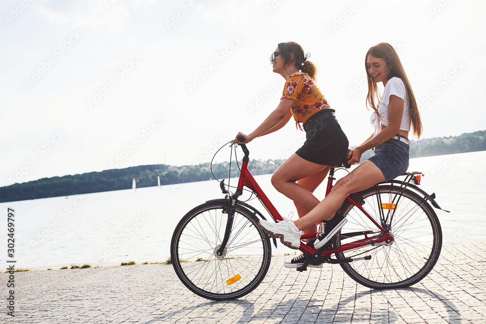 Wall mural Two female friends on the bike have fun at beach near the lake