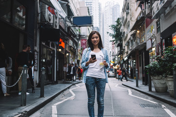 Fototapeta premium Smiling Asian woman standing on street with smartphone and drink