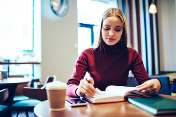 Young attractive woman blonde dressed in casual clothes planning working schedule while sitting in cozy coffee shop.Charming skilled female student preparing for examination using textbook indoors