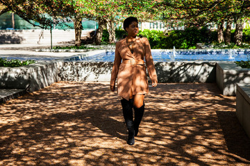 Beautiful African american  female fashion model with an Afro is walking in a park by a vibrant blue water fountain in Chicago.  the colorful serene green trees provide a peaceful summer lifestyle